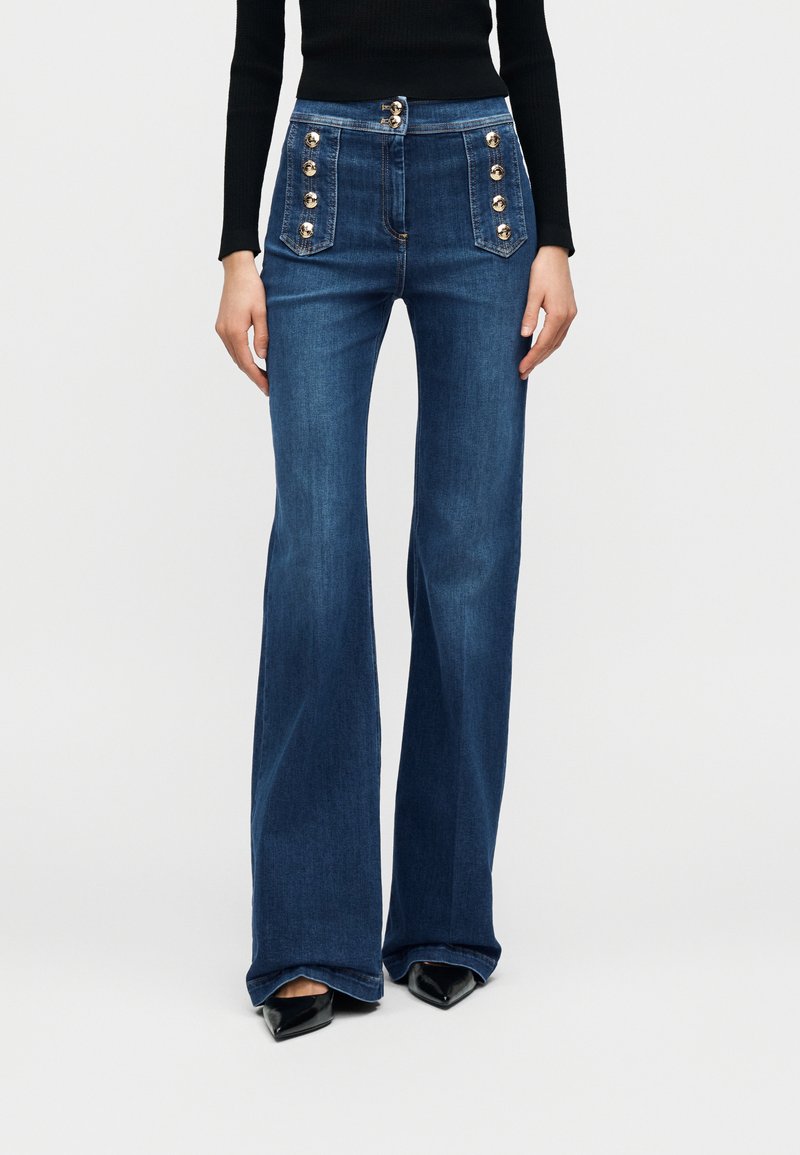 Μπλε τζιν παντελόνια με φαρδύ καμπάνα και ψηλή μέση, με διακοσμητικά χρυσά κουμπιά και μπροστινές τσέπες. Ομαλή υφή με κλασικό ξεβάμμα.