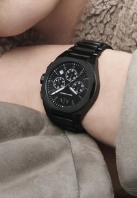 Reloj analógico negro con cara redonda, banda de acero inoxidable, múltiples subesferas y pantalla de fecha. El diseño presenta bordes afilados y angulares.