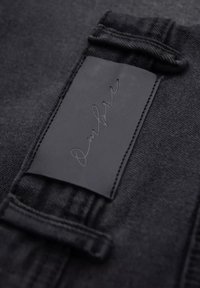 Tissu en denim noir avec une surface texturée. Une étiquette en cuir rectangulaire cousue sur la ceinture présente une écriture en relief.