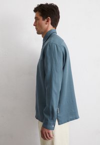 Light blue long-sleeve shirt in linen fabric, featuring a buttoned collar, chest pocket, and side slits, with subtle pink button details.