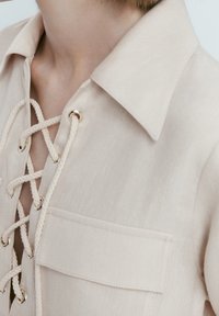 Helle beige Bluse mit Kragen und Schnürung an der Vorderseite. Verziert mit goldfarbenen Hardware-Details und einer Brusttasche. Texturiert mit einer glatten Oberfläche.