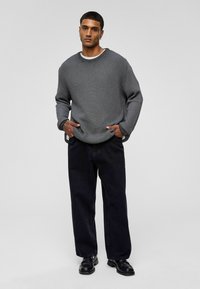 Pull en maille gris à coupe décontractée, col rond et manches longues, associé à un pantalon noir ample et des chaussures noires. Design minimaliste.