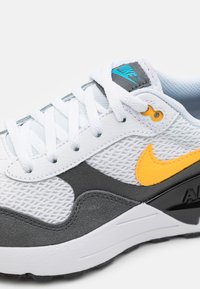 Sneaker bianca con materiali in rete e pelle sintetica. Presenta accenti azzurri, un logo Nike giallo brillante e una base grigia texturizzata.