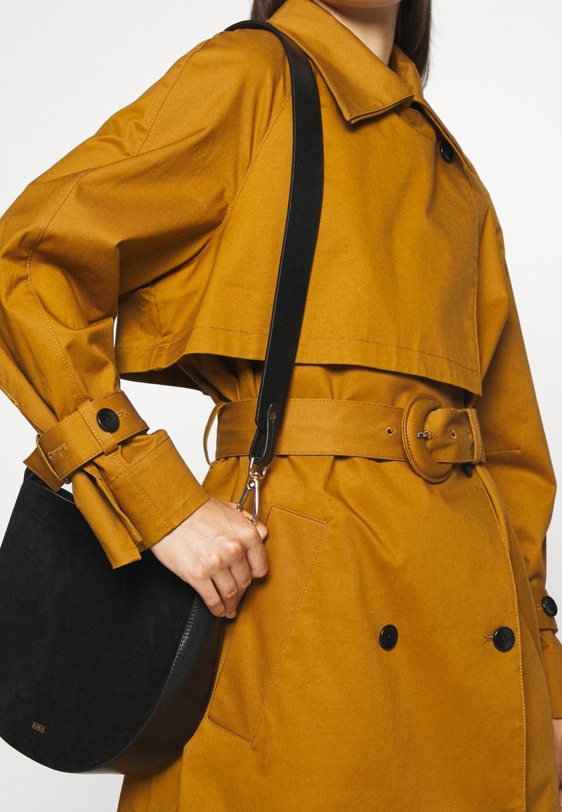 Femme portant un trench-coat moutarde ceinturé, tenant un sac à bandoulière noir avec une large sangle sur fond blanc.