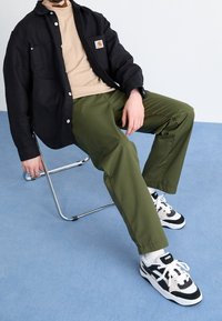 Sort skjorte med knapper og to lommer over en beige t-shirt; grønne bukser; sort og hvide sportsko; siddende på en kromstol.