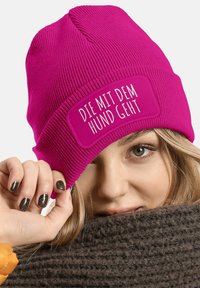 Pink knitted beanie with ribbed texture and a rectangular white patch displaying the text "DIE MIT DEM HUND GEHT." Soft fabric, fitted design.