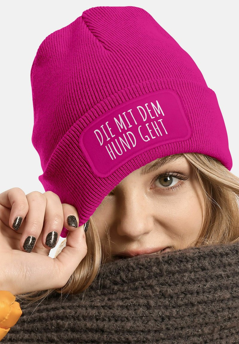 Pink knitted beanie with ribbed texture and a rectangular white patch displaying the text "DIE MIT DEM HUND GEHT." Soft fabric, fitted design.