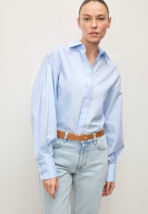 Hele sinine pikkade varrukatega nööpidega särk, millel on terav krae ja laiad varrukad, kombineeritud kõrge vöökohaga helepesuga teksapükstega ja beež vööl.