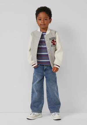 Junger Junge steht in einer grauen und cremefarbigen Collegejacke, gestreiftem Shirt, blauen Jeans und weißen Turnschuhen vor einem einfarbigen Hintergrund.