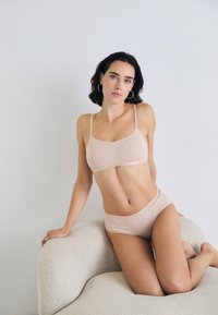 Vrouw met donker haar in huidkleurige onderkleding, knielend op een gestructureerde beige fauteuil tegen een eenvoudige witte muur.