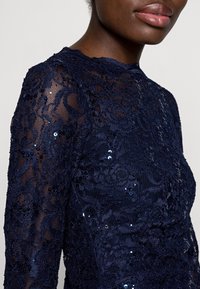 Navy lace dress with long sleeves, featuring floral patterns and sequin accents, showcasing a textured finish and a high neckline.