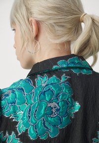 Femme blonde avec queue de cheval et boucle d'oreille argentée en forme d'anneau portant une veste noire avec une grande broderie florale turquoise et argentée.