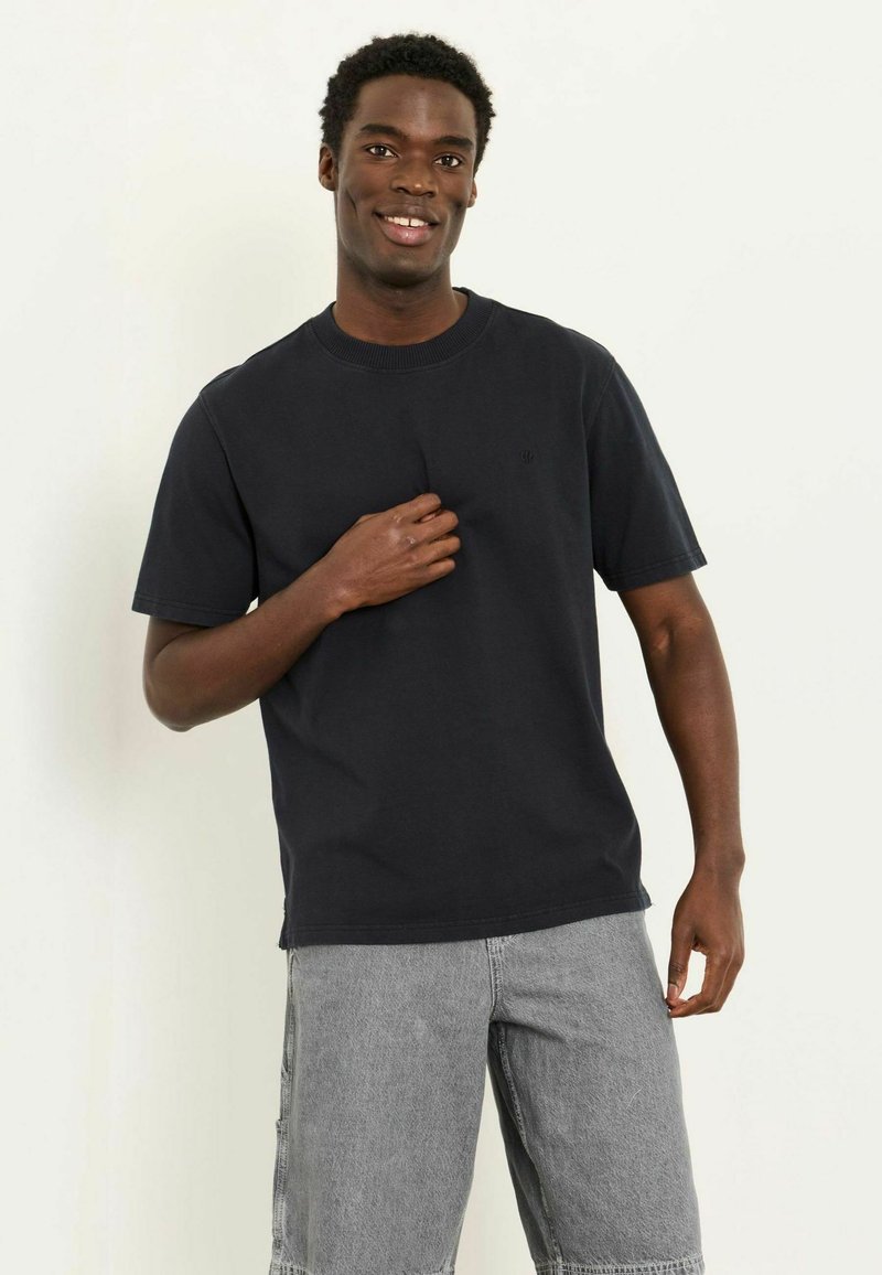 Lächelnder junger Mann, der ein schwarzes kurzärmeliges T-Shirt und graue Jeansshorts trägt, steht vor einem schlichten hellen Hintergrund.