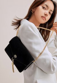 Clutch in suede nero con tracolla in catena dorata e chiusura a scatto, con dettagli a frange sul lato e flap in stile busta.