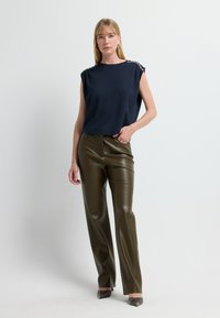 Marineblaues ärmelloses Top mit Schulterverzierungen, kombiniert mit hochtaillierten olivgrünen Kunstlederhosen und spitzen schwarzen Pumps.