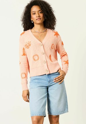 Woman with curly hair wearing a peach cardigan with orange sea creature patterns and light blue denim shorts, standing with hand on hip.