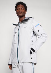 Chaqueta impermeable blanca con cremallera verde azulado, detalles en negro, múltiples bolsillos y capucha. Presenta un diseño moderno y elegante, además de un material duradero.