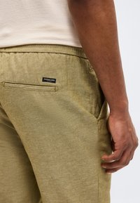 Primo piano di una persona che indossa pantaloni verde oliva con un'etichetta Scotch & Soda sulla tasca posteriore e una camicia beige a righe.