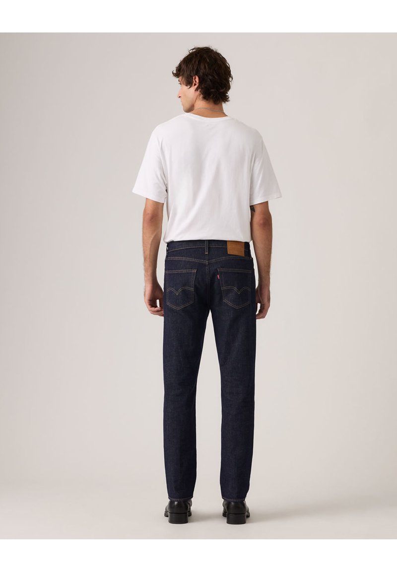 Uomo con t-shirt bianca e jeans di denim blu scuro, in piedi di spalle contro uno sfondo chiaro e uniforme.