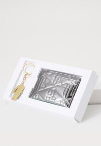 Portachiavi in oro con silhouette di figura, abbinato a un porta carte in argento texturizzato con motivi geometrici, presentato in una scatola bianca.