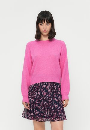 Femme portant un pull rose vif et une jupe à motifs floraux sombres se tenant devant un fond blanc uni.