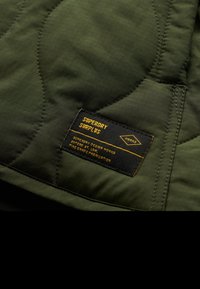 Olijfgroen, gewatteerde stof met een gestructureerd oppervlak. Zwarte label met gele tekst, met details over het merk en ontwerpinformatie.