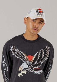 Langarm schwarzes Shirt mit einem Adler-Design und rotem Sonnen-Hintergrund. Weiße Kappe mit buntem Totenschädel- und Blumendesign.