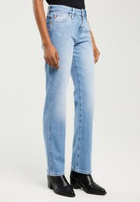 Persona, kas valkā gaiši zilas taisnas džinsa bikses, melnas potītes zābakus ar papēžiem un melnu topu, ar vienkāršu gaišu fonu.