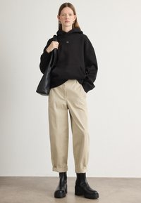 Felpa nera over-size con tasca frontale, pantaloni cropped beige e stivali platform neri. Presenta un semplice logo ricamato sulla felpa.