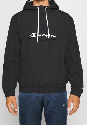 Person trägt eine schwarze Champion-Windjacke mit Kapuze, weißen Kordeln und marineblauen Champion-Trainingshosen und steht mit entspannten Armen.
