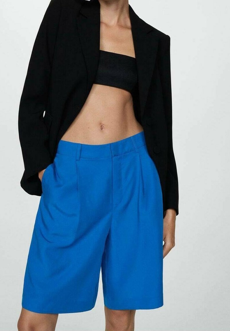 Femme portant un top court noir, un blazer noir et un short bleu ample taille haute, longueur genoux, les mains dans les poches.