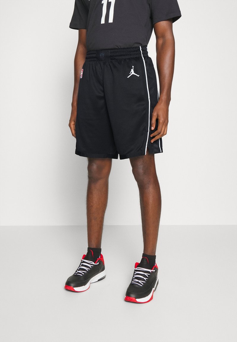 Mężczyzna w czarnych szortach do koszykówki z białymi paskami po bokach i logo Jumpman, czarnej koszulce z numerem 11 oraz czarno-czerwonych trampkach.