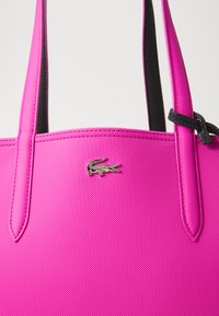 Roze synthetische schoudertas met zwarte accenten, voorzien van een gestructureerd oppervlak en een zilveren krokodillenlogo aan de voorkant. Inclusief dubbele handvatten.