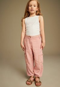 Ljust rosa cargobyxor med två sidofickor, vit ribbad linne och tan sandaler med dekorativa detaljer. Mjuk, texturerad tyg.