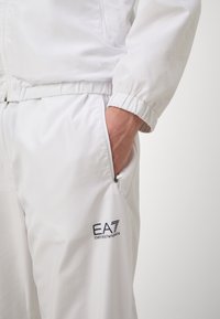 Bela vetrovka z mrežastim vzorcem, elastičnimi manšeti in z zadrgo zaprtimi stranskimi žepi. Na levem žepu je logotip EA7 Emporio Armani.
