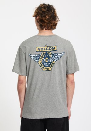 Homme aux cheveux bouclés portant un t-shirt gris avec un logo Volcom ailé et le texte « Stone Co. CA Since 1991 True to This » au dos.