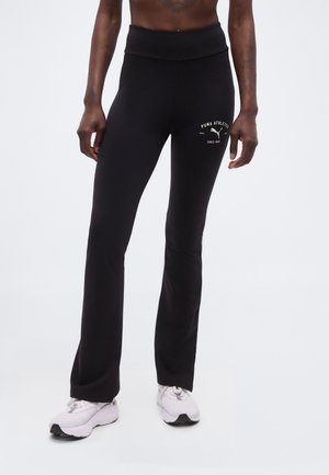 Osoba má na sebe čierne legíny Puma Athletics s bielym logom na stehne a biele športové topánky, stojí na svetlom pozadí.