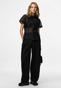 Blouse noire transparente à manches courtes avec boutons devant et col, associée à un pantalon noir ample taille haute et des chaussures noires texturées.