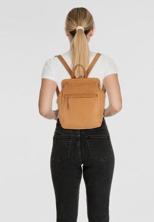 Zaino in pelle marrone con forma rettangolare, tasca frontale con zip e spallacci regolabili. Texture liscia, design minimalista e manico superiore.
