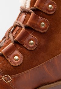 Bota de invierno marrón de cuero y ante con ojales de metal, cordones gruesos y forro de suave forro polar visible.