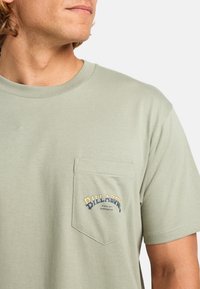 Lys grøn bomuld t-shirt med en brystlomme med blå og guld skrift. Rund halsudskæring med korte ærmer. Glat tekstur.
