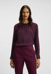 Langärmlige Bluse in tiefem Burgunderrot, mit gerafftetem Ausschnitt und glattem Stoff. Kombiniert mit passender, maßgeschneiderter Hose für einen einheitlichen Look.