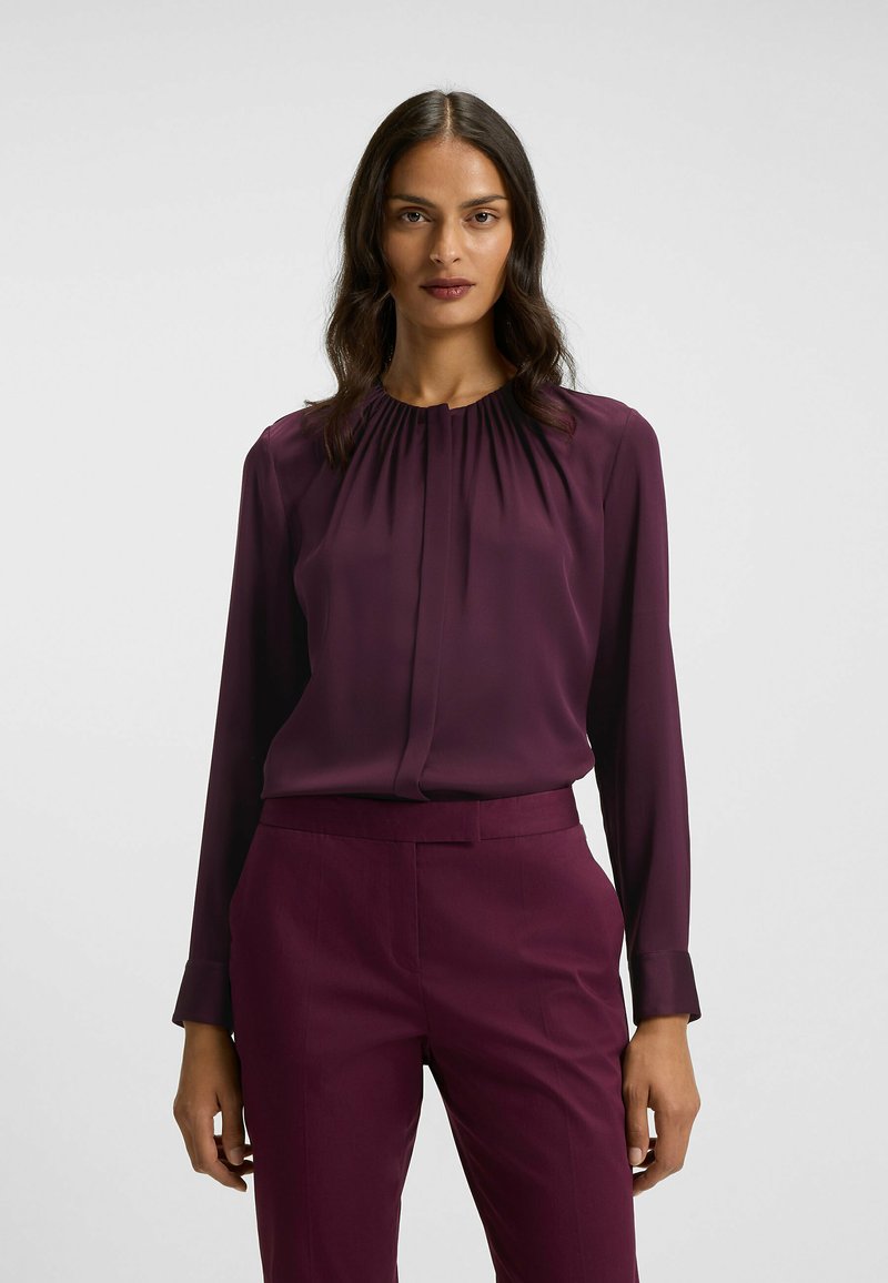 Langärmlige Bluse in tiefem Burgunderrot, mit gerafftetem Ausschnitt und glattem Stoff. Kombiniert mit passender, maßgeschneiderter Hose für einen einheitlichen Look.
