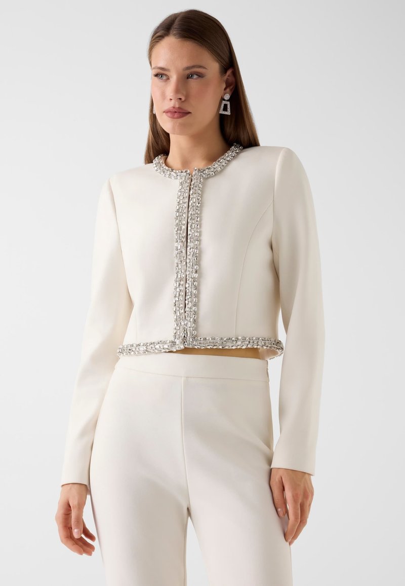 Mujer con conjunto de chaqueta corta y pantalones en color blanco roto, con ribete decorativo en los bordes de la chaqueta y pendientes a juego.