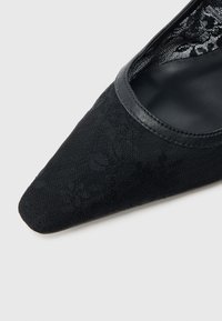 Μαύρο παπούτσι με μυτερή μύτη από υφασμάτινη υφή με ανθικά μοτίβα και γυαλιστερό μαύρο περίγραμμα κατά μήκος του ανοίγματος. Το εσωτερικό είναι λείο.