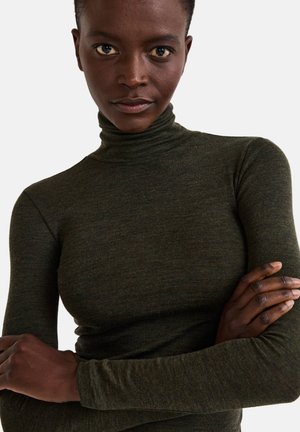 Personne confiante avec une peau foncée et des cheveux courts, portant un pull à col roulé vert olive, les bras croisés devant un fond uni.