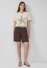 Beige t-shirt met korte mouwen met een kleurrijk zeester ontwerp, gecombineerd met donkerbruine opgerolde shorts en bruine sandalen met een gestreepte band.