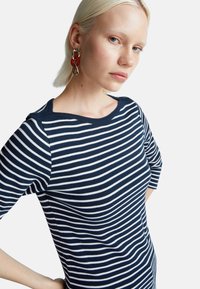 Niebiesko-biała koszula w paski z okrągłym dekoltem i rękawami o długości trzy czwarte, uszyta z miękkiego materiału. Modelka nosi wyraziste kolczyki w kolorze czerwonym i złotym.