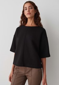 Top corto nero a maniche corte con scollo ampio e vestibilità morbida. Realizzato in materiale soffice, presenta cuciture a vista ed è abbinato a pantaloni marroni aderenti.