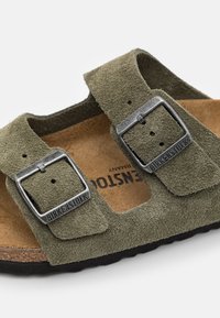Olivgrön mockasandal med två justerbara remmar och silver spännen. Brun fotsäng med logotyp, strukturerad svart sula.